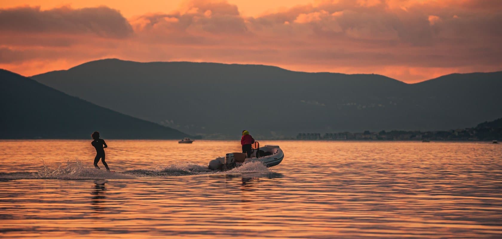 A boat towing a waterskier across the Bay of Tivat at sunset, with the mountains behind